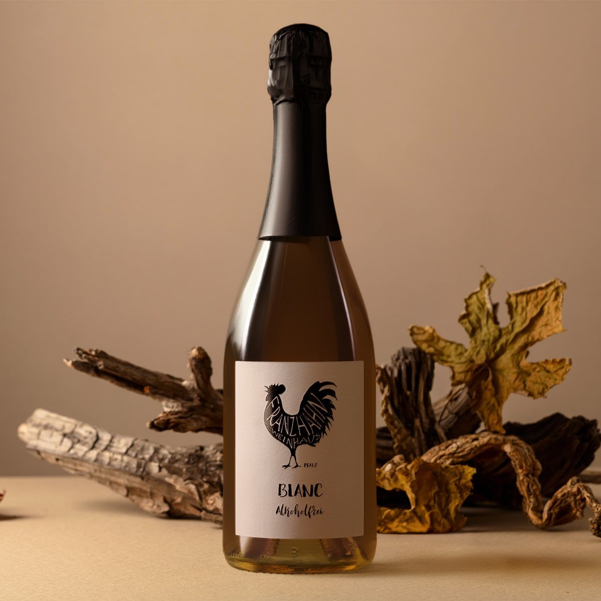 Eine Sektflasche mit der Aufschrift "Blanc" und dem Logo eines schwarzen Hahns steht auf einer beigen Fläche, umgeben von getrockneten Blättern und Holzstücken vor einem neutralen Hintergrund.