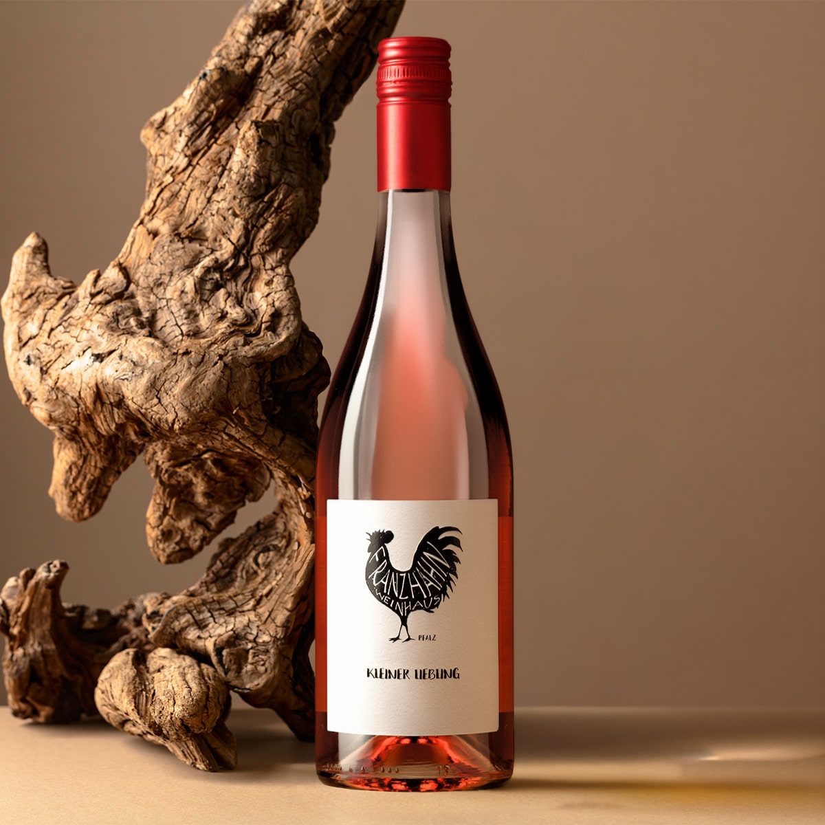 Eine Flasche Roséwein mit rotem Verschluss und einem Etikett mit schwarzem Hahnmotiv steht neben einem Stück gedrehten Treibholzes auf einer beigen Fläche mit neutralem Hintergrund.