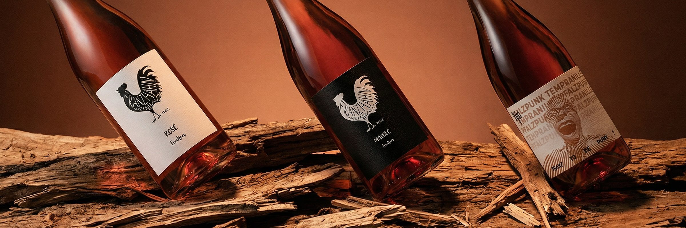 © Weinhaus Franz Hahn Drei Flaschen Roséwein mit Hahn-Etiketten sind schräg auf rustikalen Holzteilen vor einem warmen braunen Hintergrund ausgestellt.