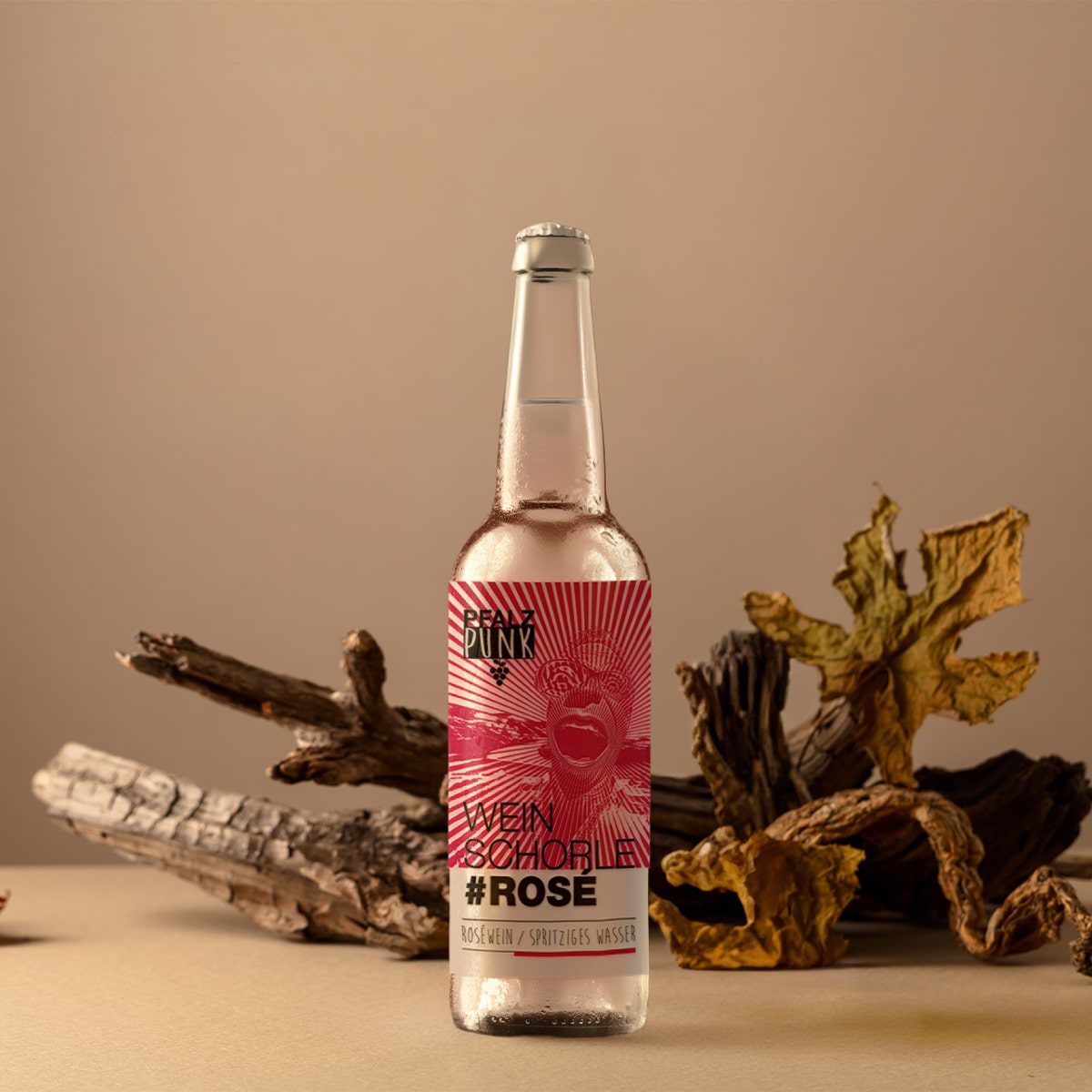 Eine Klarglasflasche mit der Aufschrift "Schorle Rosé" steht aufrecht auf einer beigen Fläche, im Hintergrund sind Treibholz und getrocknete Blätter vor einem neutralen Hintergrund angeordnet.