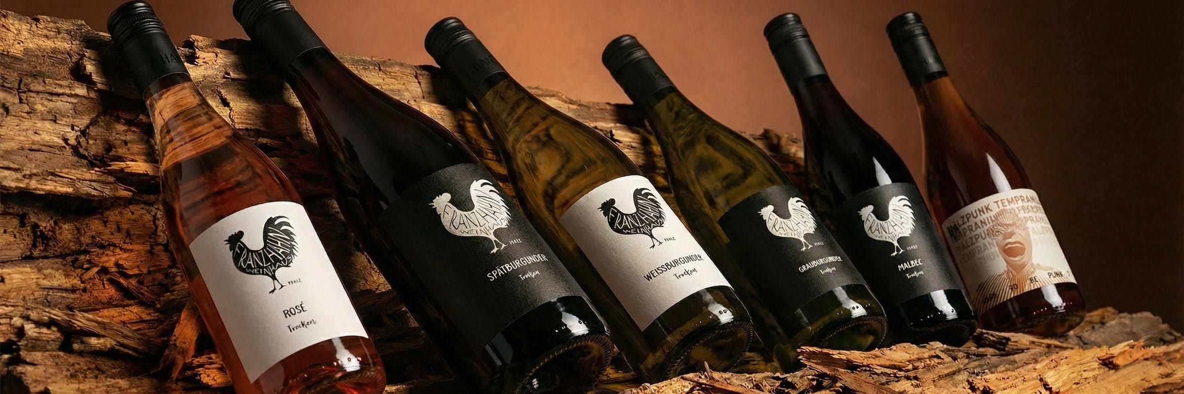 © Weinhaus Franz Hahn Sechs Weinflaschen mit schwarzen Hahn-Etiketten sind auf rauen Holzstämmen ausgestellt. Bei den Flaschen handelt es sich um Rosé-, Weiß- und Rotweine, die schräg vor einem warmen, braunen Hintergrund stehen.
